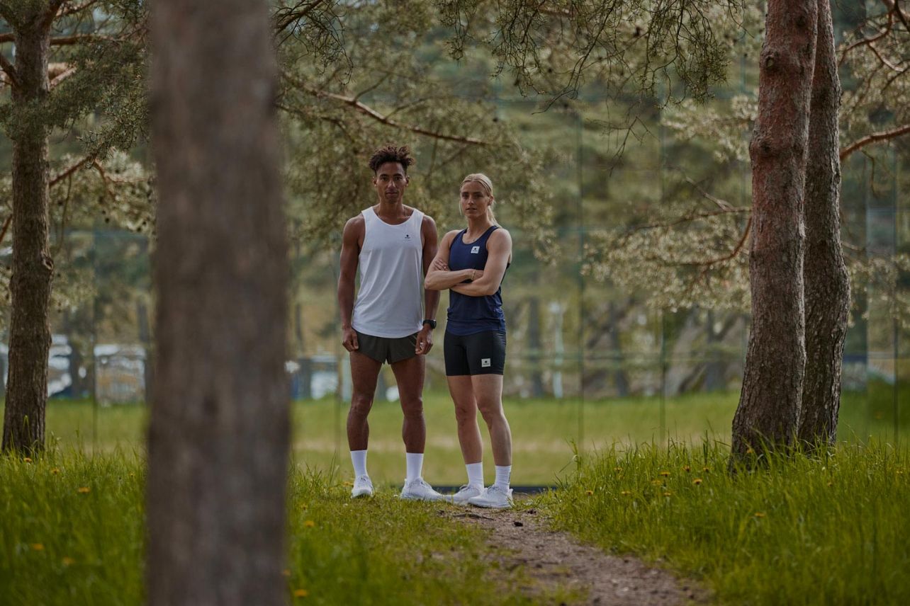 To løpere poserer i teknisk løpetøy fra Saysky mellom trær i skogen.