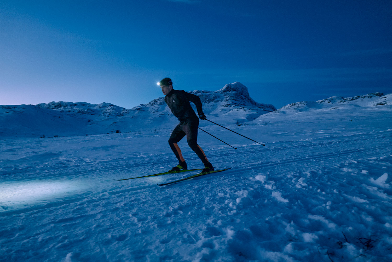 Mann på langrennstur i vinterfjellet med hodelykt i skumringen.