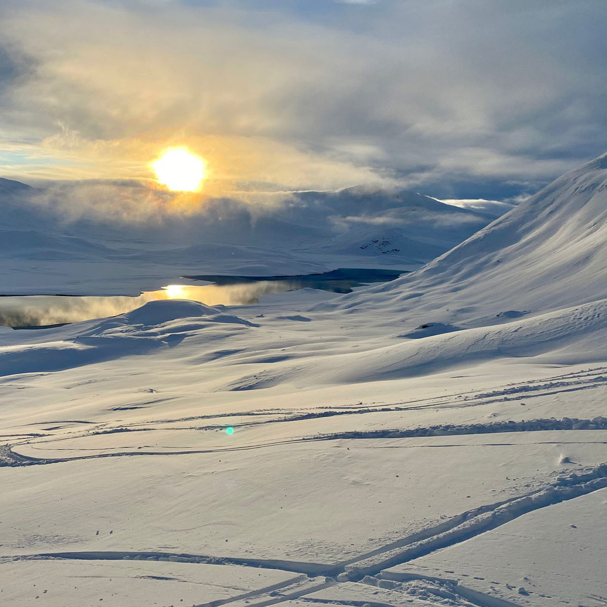 Snødekt fjellandskap med skispor i forgrunnen og lav vintersol over horisonten.