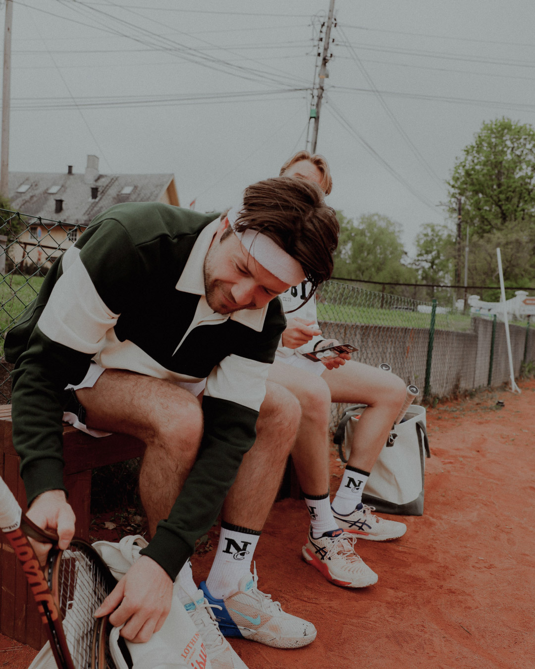 To menn ikledd Nibbi Clothing tennisantrekk gjør seg klare til kamp på grusbane. Den ene pakker ut racketen, den andre sjekker mobilen.